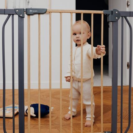 Batoľa stojí za drevenou bezpečnostnou zábranou s kovovými bočnými časťami v interiéri. Na podlahe sú hračky. Na zábranu je pripevnená značka s nápisom Lionelo.