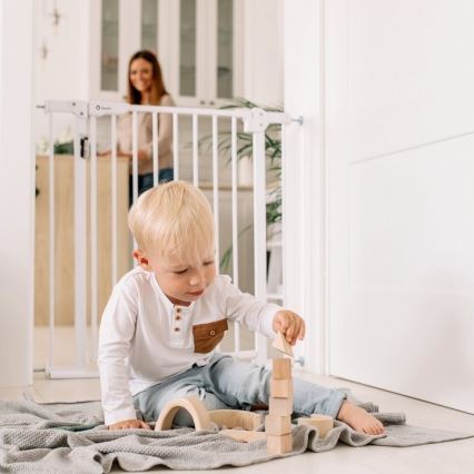Dieťa stavia drevené kocky na podlahe. V pozadí je bezpečnostná bránka a žena. Dieťa sedí na svetlošedej deke.