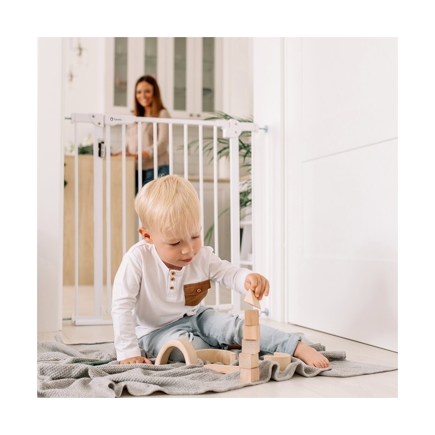 Dieťa stavia drevené kocky na podlahe. Na pozadí je bezpečnostná bránička a žena.