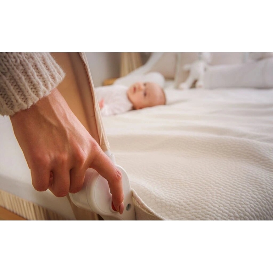 Ruka nastavuje bezpečnostnú zábranu detskej postieľky. Dieťa leží v pozadí na posteli. Zábrana je béžová a biela, posteľ je svetlo béžová.