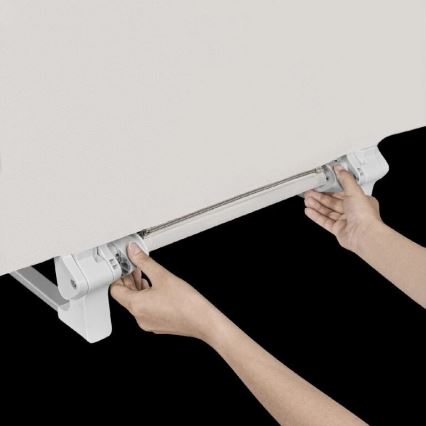 Dve ruky ovládajú posúvač látkovej rolety pripevnený k bielemu mechanizmu. Roleta je svetlo béžová a zasunutá do bieleho rámu.