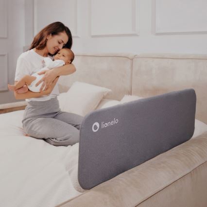 Žena drží dojča na posteli s ochrannou bariérou. Bariéra je sivá, s logom „lionelo“. Posteľ je svetlá, v spálni.