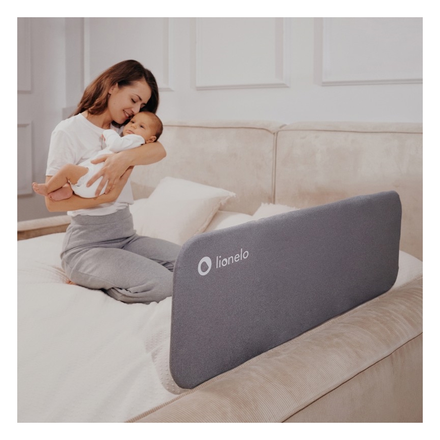 Žena drží dojča na posteli s ochrannou bariérou. Bariéra je sivá, s logom „lionelo“. Posteľ je svetlá, v spálni.