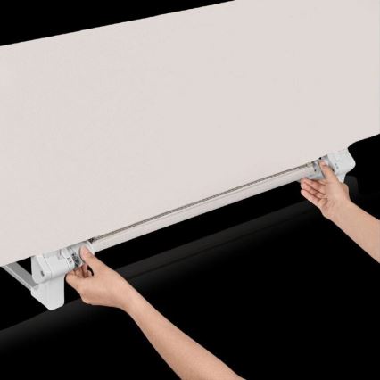 Dve ruky ovládajú mechanizmus rolety. Svetlo béžová látka rolety sa pohybuje po koľajničke. Koľajnička je pripevnená k bielemu držiaku.