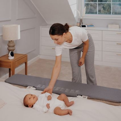 Dojča leží na sivej podložke na posteli. Matka sa k nemu skláňa. V pozadí je nočný stolík s lampou a bezpečnostnou kamerou.