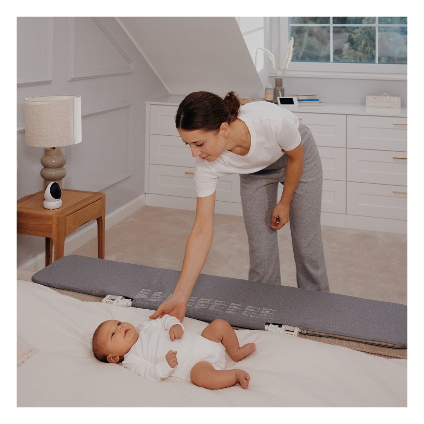 Dojča leží na sivej podložke na posteli. Matka sa k nemu skláňa. V pozadí je nočný stolík s lampou a bezpečnostnou kamerou.