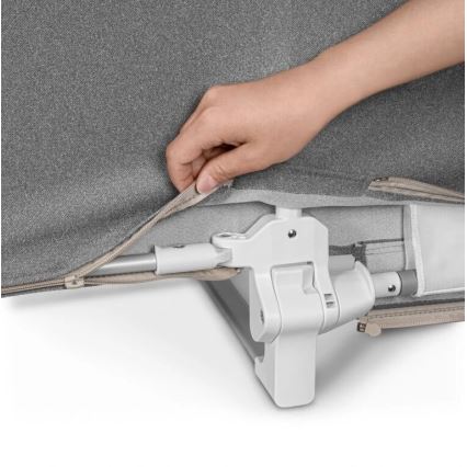 Ruka zapína zips sivého poťahu na detskej postieľke s bielym mechanizmom. Poťah je prichytený k kovovej konštrukcii postieľky.