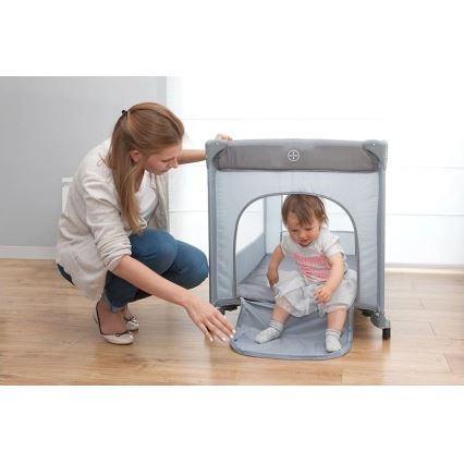 Dieťa sedí v prenosnej postieľke s mäkkým dnom a otváracím vchodom. Matka pri nej kľačí a upravuje vstup. Postieľka stojí na drevenej podlahe v svetlej miestnosti.