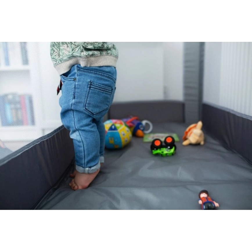 Dieťa v džínsoch stojí v detskej ohrádke. Na podlahe ohrádky sú roztrúsené hračky: lopta, autíčka a plyšová hračka. Ohrádka je sivá a má sieťové steny.