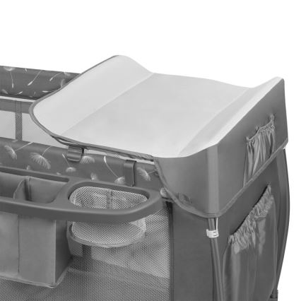 Prebaľovacia podložka je umiestnená na vrchu prenosnej postieľky. Postieľka je sivá s obrázkami púpavy, obsahuje bočné vrecká a odkladacie priestory.