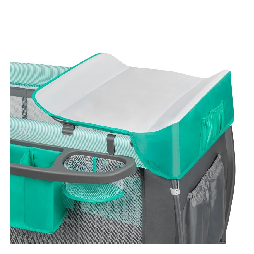 Prebaľovací pult je umiestnený na detskej cestovnej postieľke. Postieľka má sivú konštrukciu, tyrkysové prvky a sieťované vrecká. Pult je biely s tyrkysovým lemom.