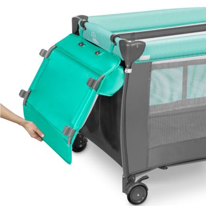 Prebaľovací pult sa pripája k cestovnej postieľke. Postieľka je šedo-tyrkysovej farby, má kolieska a sieťované steny. Ruka pripája pult k postieľke.