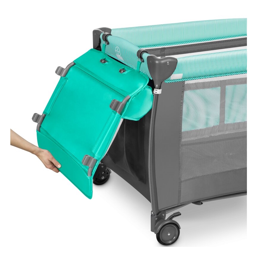 Prebaľovací pult sa pripája k cestovnej postieľke. Postieľka je šedo-tyrkysovej farby, má kolieska a sieťované steny. Ruka pripája pult k postieľke.