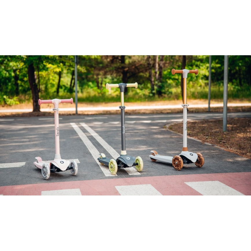 Tri detské odrážadlá stoja na chodníku. Každé má tri kolesá a inú farbu: ružovú, sivú a svetlo béžovú. Na rukovätiach je nápis „Lineolo“. Za odrážadlami je zeleň stromov.