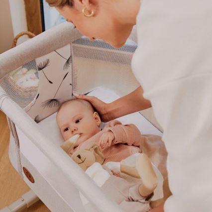 Dojča leží v prenosnej postieľke s motívom púpavy. Žena sa nad ním skláňa. Postieľka je svetlo šedá s bielym textilným vnútrom.