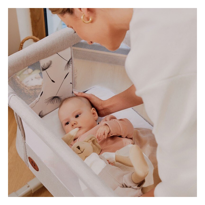 Dojča leží v prenosnej postieľke s motívom púpavy. Žena sa nad ním skláňa. Postieľka je svetlo šedá s bielym textilným vnútrom.