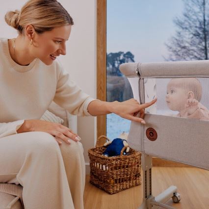 Žena ukazuje na prenosnú postieľku s dojčaťom vnútri. Postieľka je béžová, s oknom zo sieťoviny. Nachádza sa v svetlej izbe s drevenou podlahou a košíkom hračiek.