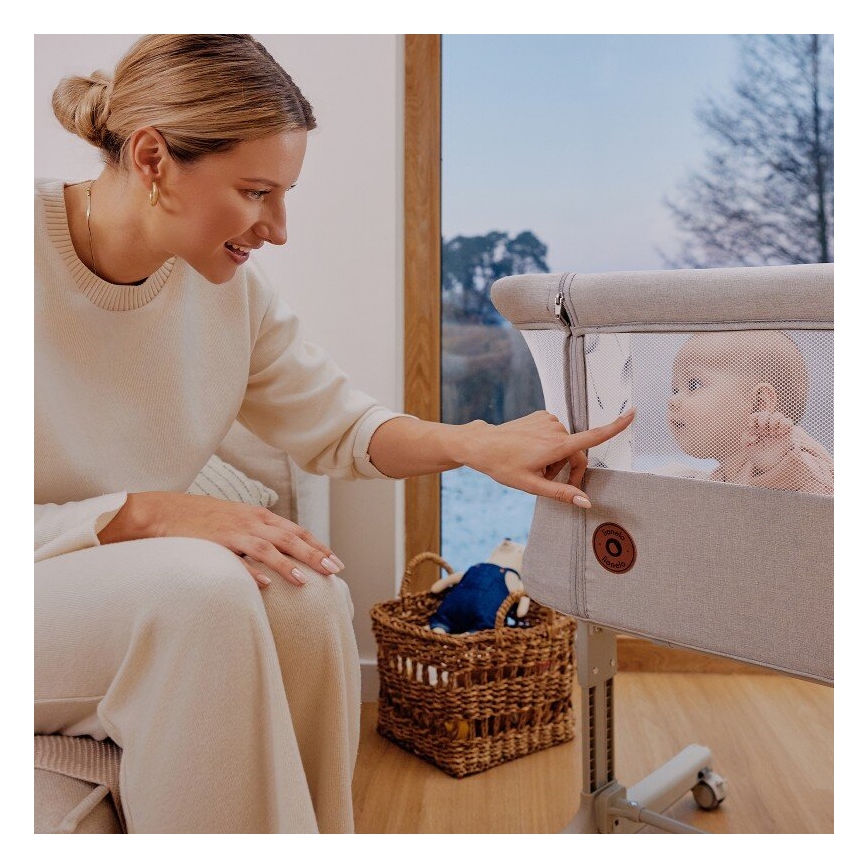 Žena ukazuje na prenosnú postieľku s dojčaťom vnútri. Postieľka je béžová, s oknom zo sieťoviny. Nachádza sa v svetlej izbe s drevenou podlahou a košíkom hračiek.