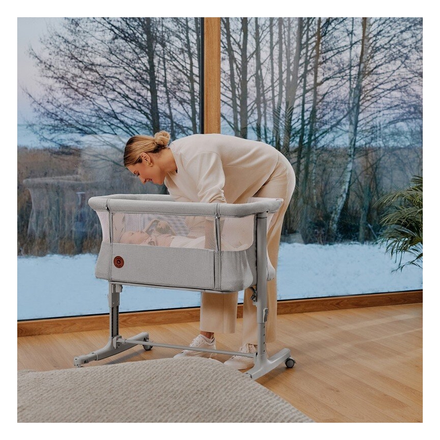 Žena sa nakláňa nad šedou prenosnou postieľkou s bábätkom. Postieľka stojí na kolieskach v svetlej izbe s výhľadom na zasneženú krajinu za veľkým oknom.