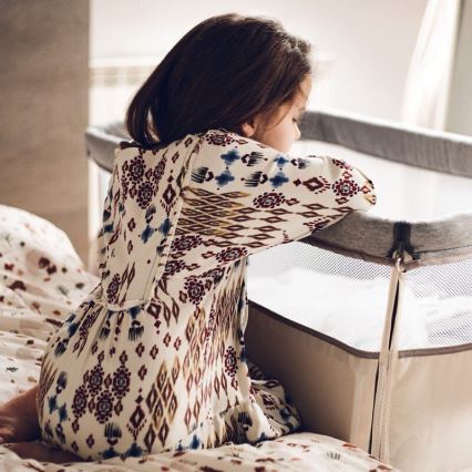 Dievča v šatách s kvetinovým vzorom sedí na posteli a opiera sa o prenosnú postieľku. Postieľka je béžová s šedým lemom a sieťovanými stranami. Dievča sa pozerá dole.