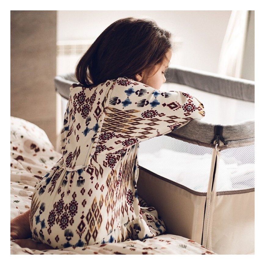 Dievča v šatách s kvetinovým vzorom sedí na posteli a opiera sa o prenosnú postieľku. Postieľka je béžová s šedým lemom a sieťovanými stranami. Dievča sa pozerá dole.