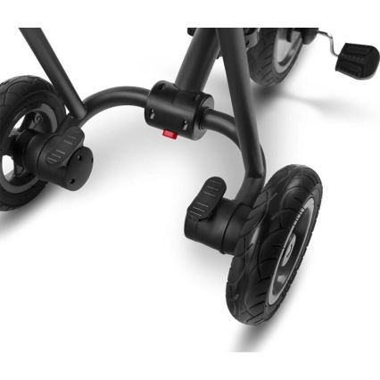 Trojkolka s otočnými kolesami a pedálmi je zachytená z nízkeho uhla. Konštrukcia je prevažne čierna, s červeným detailom na spojovacom mechanizme. Viditeľné sú gumové pneumatiky a otočné mechanizmy na kolesách.