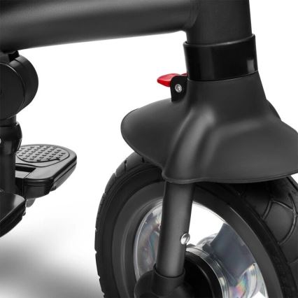 Predné koleso trojkolky s blatníkom a červenou páčkou je zobrazené v detailnom zábere. Koleso je čierne, s gumenou pneumatikou. Na ráme je viditeľná opierka pre nohy.