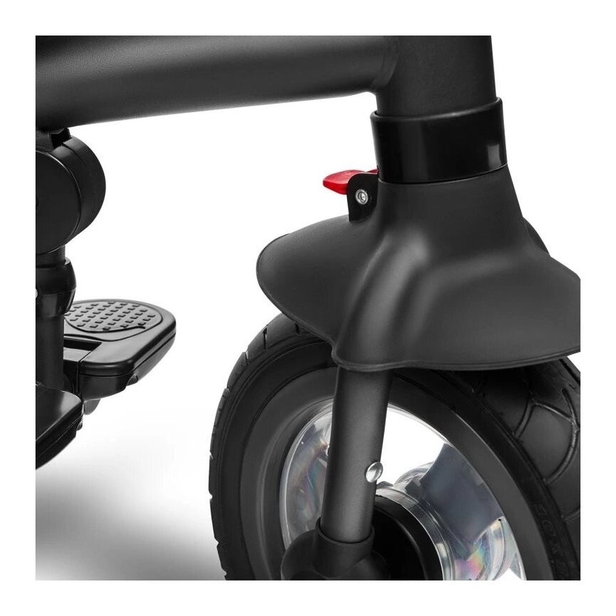 Predné koleso trojkolky s blatníkom a červenou páčkou je zobrazené v detailnom zábere. Koleso je čierne, s gumenou pneumatikou. Na ráme je viditeľná opierka pre nohy.