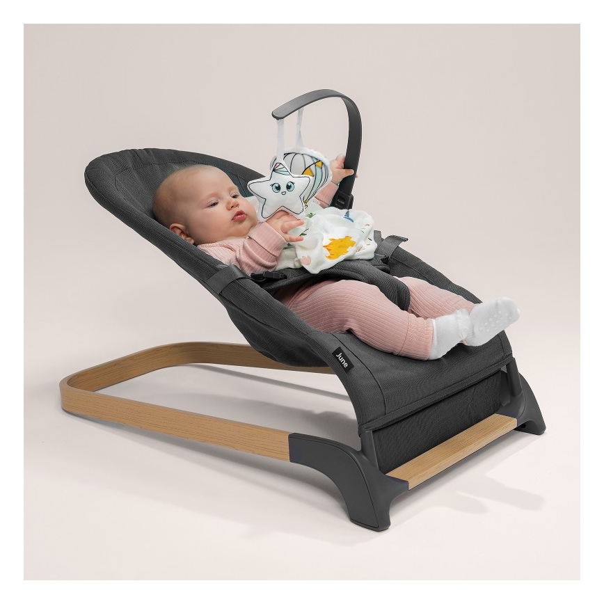 Dojča leží v šedej ležiacej sedačke s dreveným rámom a hrá sa s pripevnenou hrazdičkou s plyšovou hviezdou. Sedačka je tmavošedá, má značku Jane.