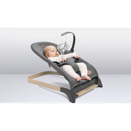 Dojča leží v sivej ležadle s drevenými nožičkami a hrazdičkou s plyšovou hračkou v tvare balónu. Ležadlo je umiestnené na svetlej ploche.