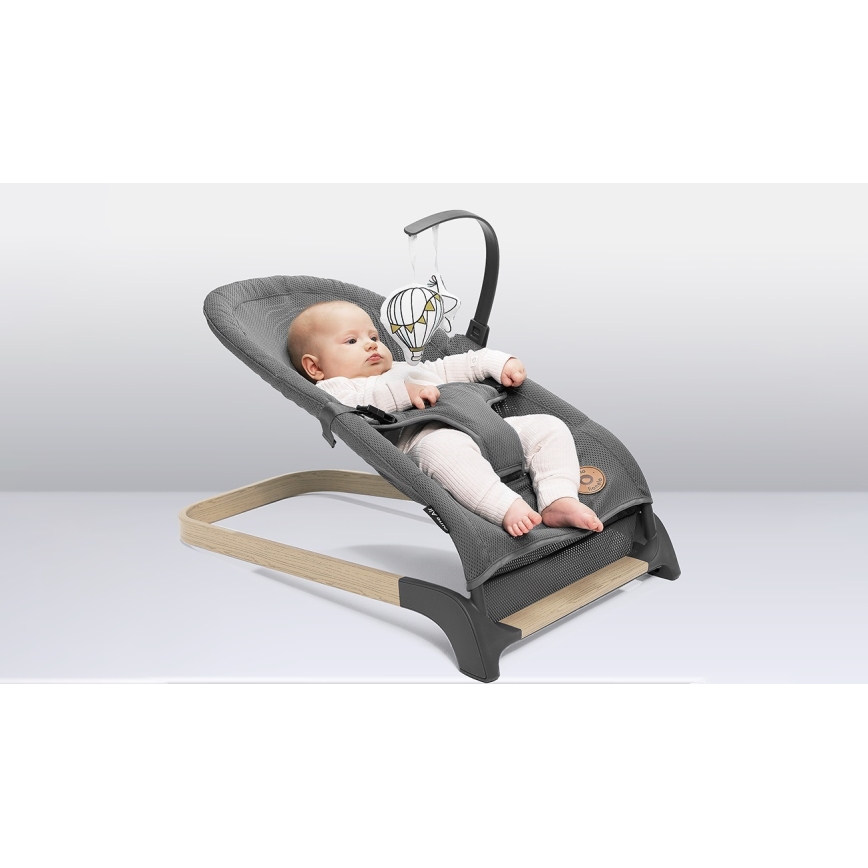 Dojča leží v sivej ležadle s drevenými nožičkami a hrazdičkou s plyšovou hračkou v tvare balónu. Ležadlo je umiestnené na svetlej ploche.