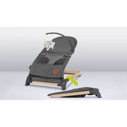 Šedá ležadlová sedačka pre bábätká s textilnou sieťovinou a dreveným rámom, je zobrazená s prídavnou hračkou. Sedačka je položená na podlahe, jej podnožka je odnímateľná a leží vedľa.