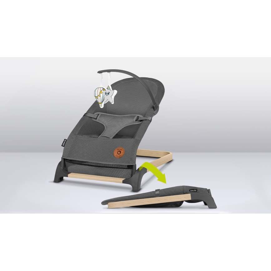 Šedá ležadlová sedačka pre bábätká s textilnou sieťovinou a dreveným rámom, je zobrazená s prídavnou hračkou. Sedačka je položená na podlahe, jej podnožka je odnímateľná a leží vedľa.