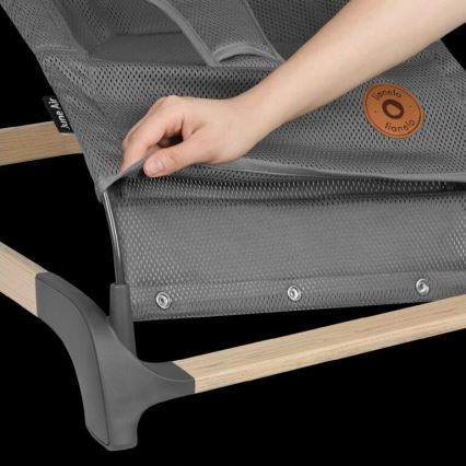 Ruka napína sivú sieťovinu lehátka s drevenými a šedými časťami konštrukcie. Na sieťovine je viditeľné logo „Lionelo“. Lehátko je položené na tmavom pozadí.