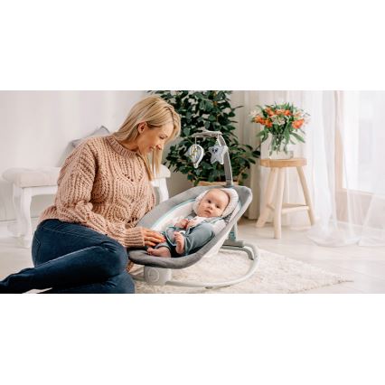 Dojča leží v hojdačke, ktorú drží matka. Hojdačka je sivá a svetlomodrá, má nad sebou mobil s hračkami. Kontext tvorí svetlá obývacia izba.