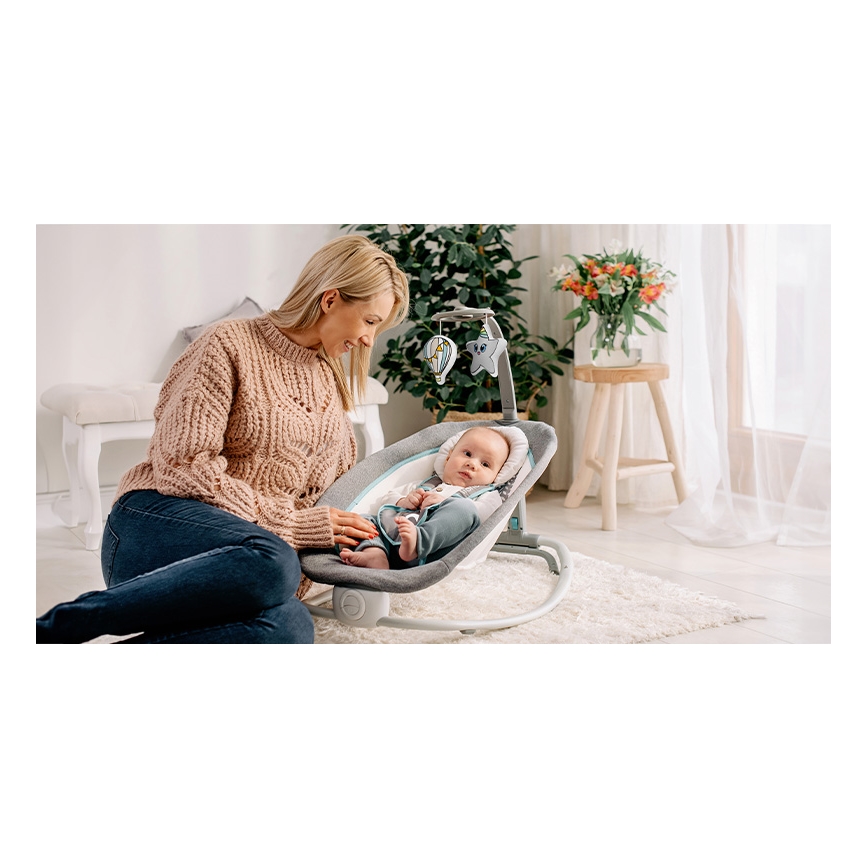 Dojča leží v hojdačke, ktorú drží matka. Hojdačka je sivá a svetlomodrá, má nad sebou mobil s hračkami. Kontext tvorí svetlá obývacia izba.