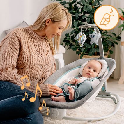 Dojča leží v hojdacej sedačke s hračkami nad hlavou. Matka sedí vedľa a usmieva sa na neho. Sedačka je v obývacej izbe.