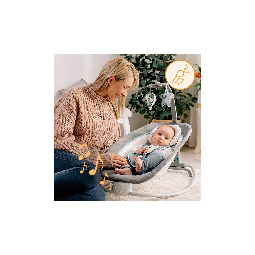 Dojča leží v hojdacej sedačke s hračkami nad hlavou. Matka sedí vedľa a usmieva sa na neho. Sedačka je v obývacej izbe.