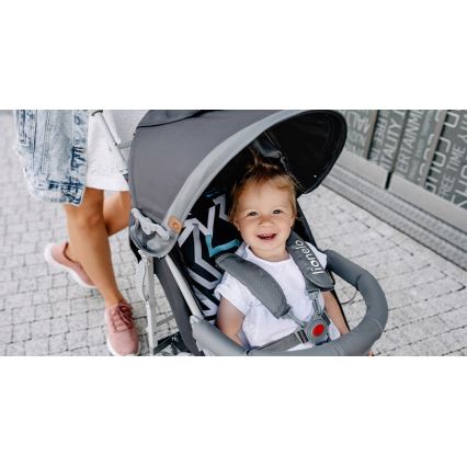 Dieťa sedí v športovom kočíku Lionelo. Kočík je tmavosivý s prekrytím proti slnku. Dieťa sa usmieva. Matka ide vedľa kočíka po dlažbe.