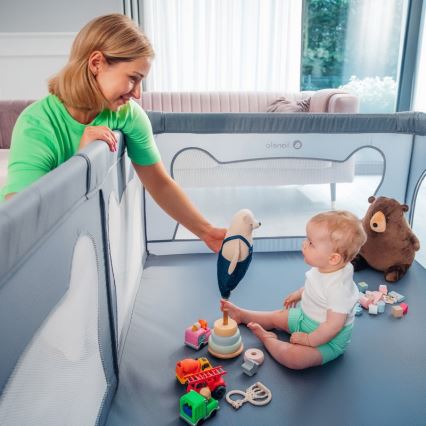 Dieťa sedí v ohrádke s hračkami. Žena pri ohrádke drží hračku. Ohrádka je šedá s bielymi sieťkami.