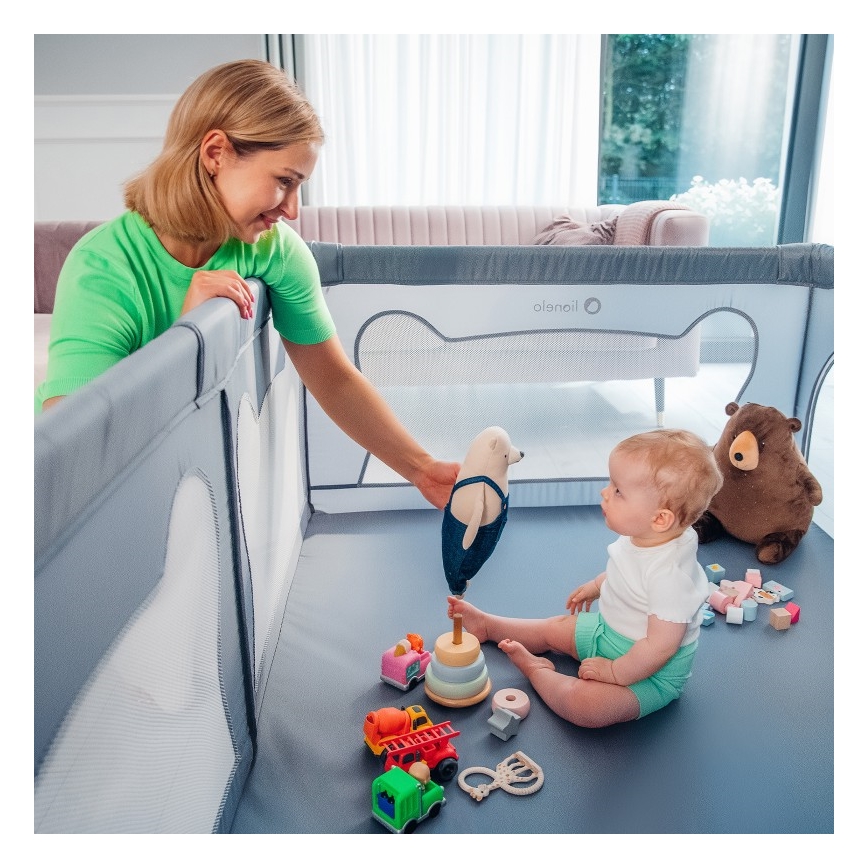 Dieťa sedí v ohrádke s hračkami. Žena pri ohrádke drží hračku. Ohrádka je šedá s bielymi sieťkami.