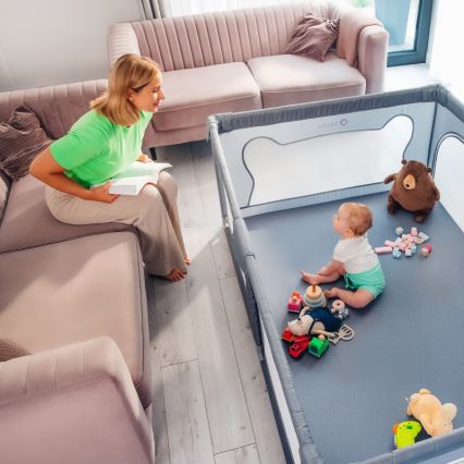 Žena sedí na pohovke s knihou, zatiaľ čo malé dieťa sedí v ohrádke s hračkami. Ohrádka je umiestnená na podlahe v obývacej izbe.