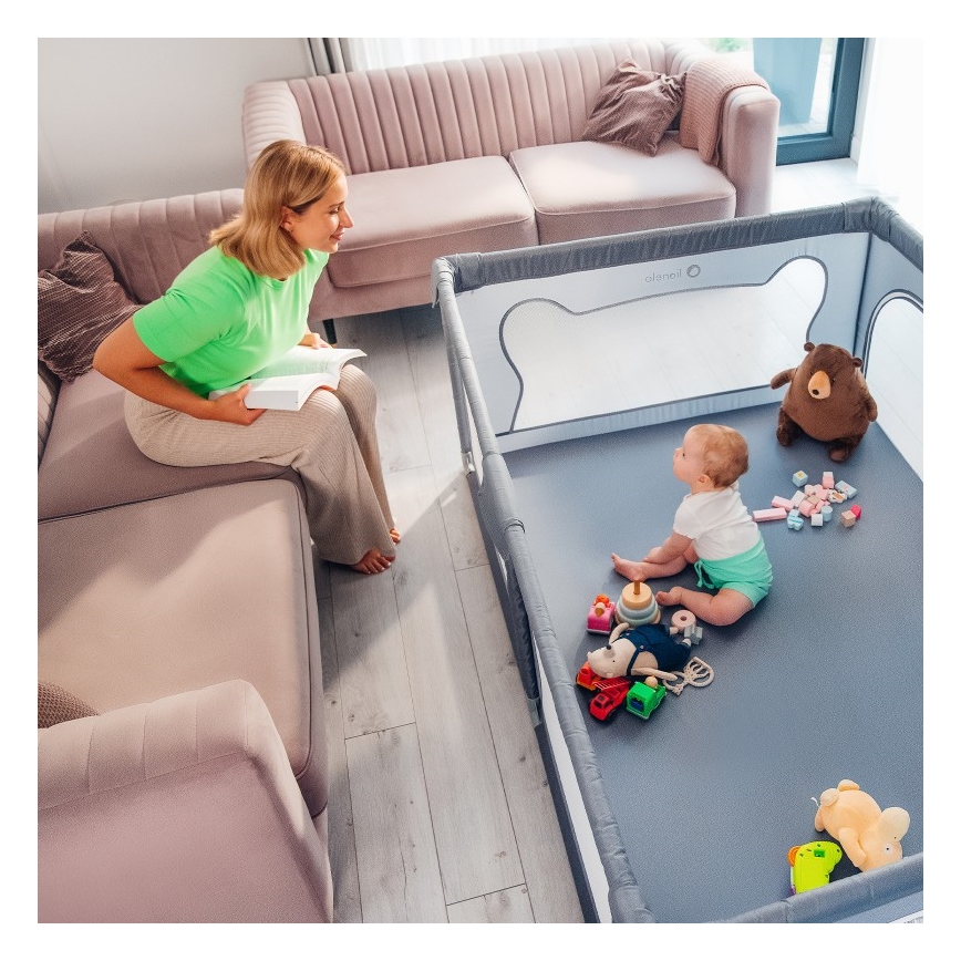 Žena sedí na pohovke s knihou, zatiaľ čo malé dieťa sedí v ohrádke s hračkami. Ohrádka je umiestnená na podlahe v obývacej izbe.