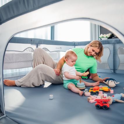 Dieťa si hrá s hračkami v ohrádke. Matka sedí vedľa neho a sleduje ho. Ohrádka je v svetlej miestnosti.