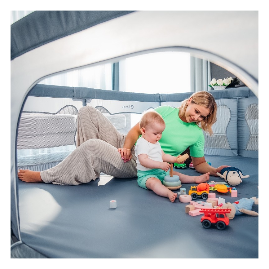 Dieťa si hrá s hračkami v ohrádke. Matka sedí vedľa neho a sleduje ho. Ohrádka je v svetlej miestnosti.