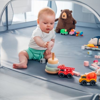 Dieťa hrá s drevenou pyramídou a autíčkami v ohrádke. Bábätko sedí na mäkkej podložke a manipuluje s farebnými hračkami. V pozadí je plyšový medvedík a ďalšie hračky.