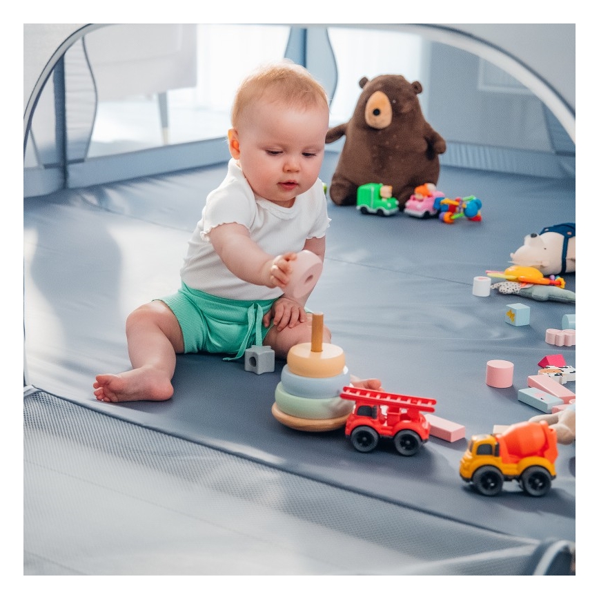 Dieťa hrá s drevenou pyramídou a autíčkami v ohrádke. Bábätko sedí na mäkkej podložke a manipuluje s farebnými hračkami. V pozadí je plyšový medvedík a ďalšie hračky.