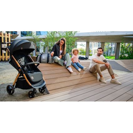 Kočík stojí na drevenej ploche. Rodina s malým dieťaťom sedí na blízku. Na taške je nápis BABY.