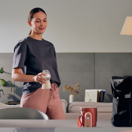 Žena drží fľašku s mliekom v obývacej izbe. Na stole stojí ohrievač fliaš. Vedľa je taška.