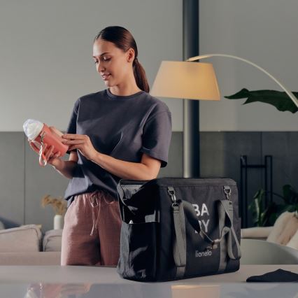 Žena kontroluje cumlík v ružovom obale. Drží ho pri čiernej taške na prenášanie s nápisom „BABY“ a značkou „lionelo“. Je v obývačke.